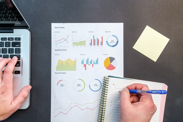 laptop, data charts, and a notebook being written in on a desk